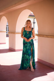 Calypso Tropical Maxi Dress with a vibrant green print, keyhole detail, and a side slit, walking under an arched terrace.