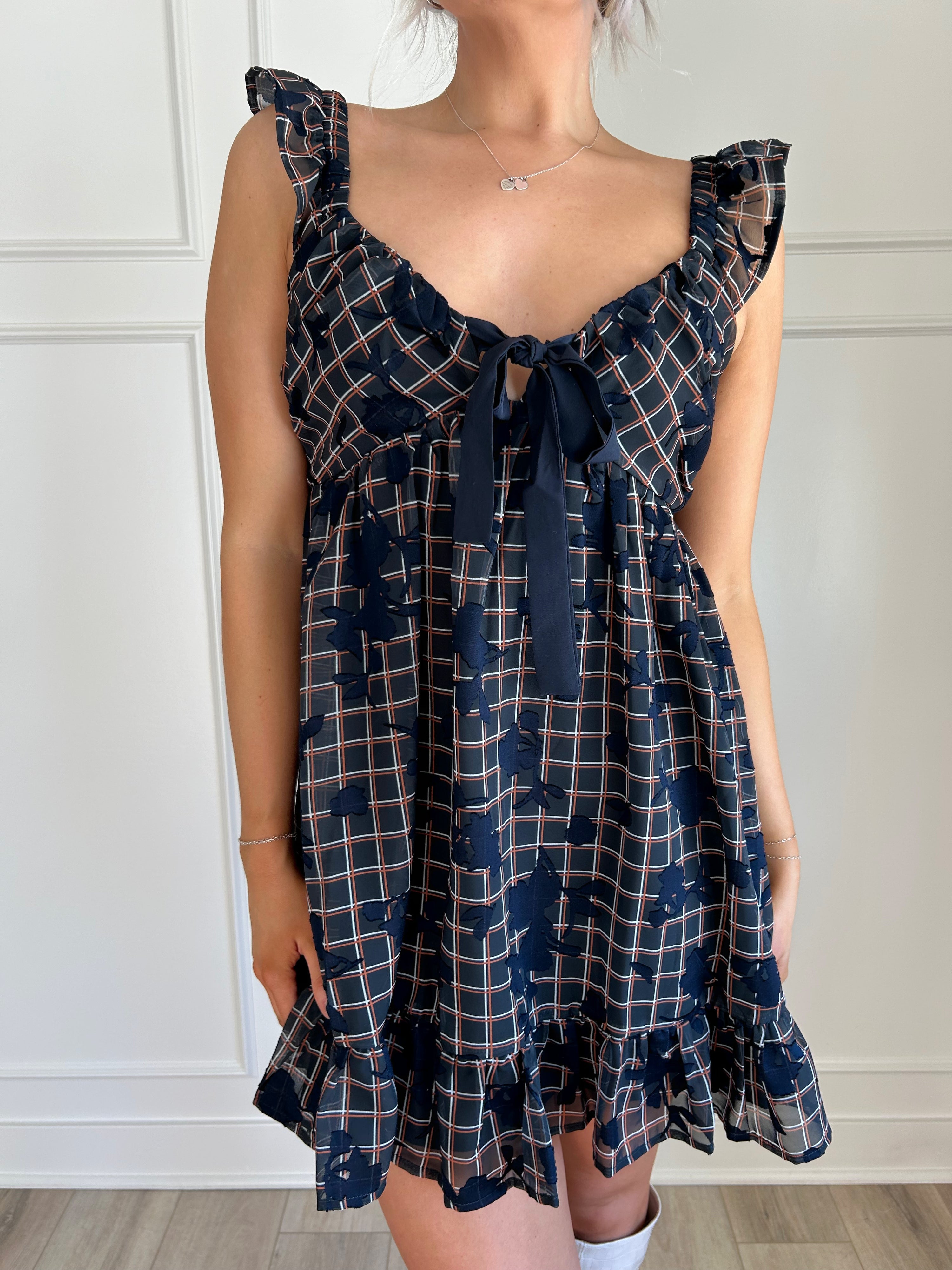 Navy plaid babydoll dress with ruffle sleeves, bow tie detail, and playful hemline.